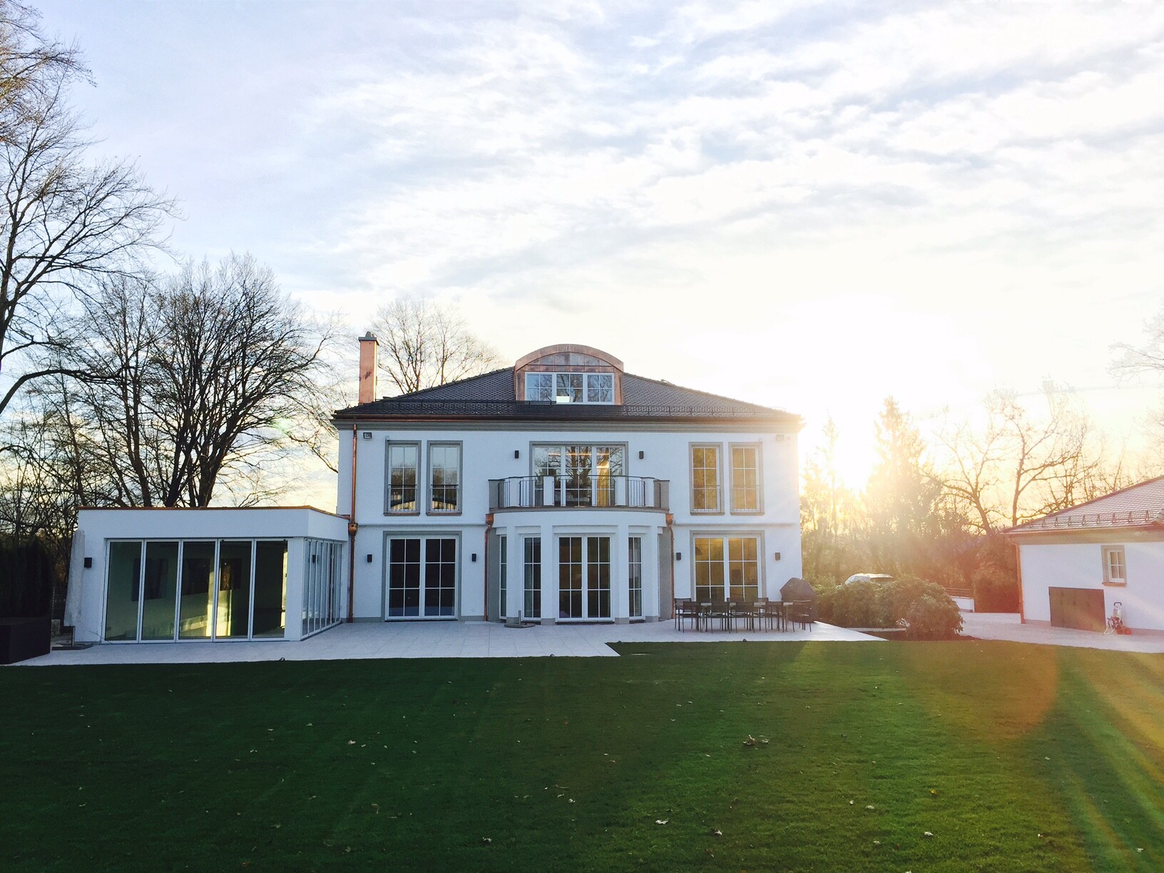 Weiße Villa mit linksseitigem Glasanbau, Balkon, Terrasse und grünem Rasen bei Sonnenuntergang.