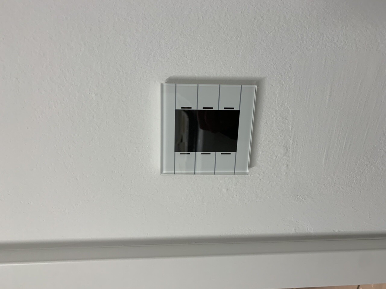 Weißes, quadratisches Wandgerät mit kleinem schwarzem Bildschirm an grauer Wand.