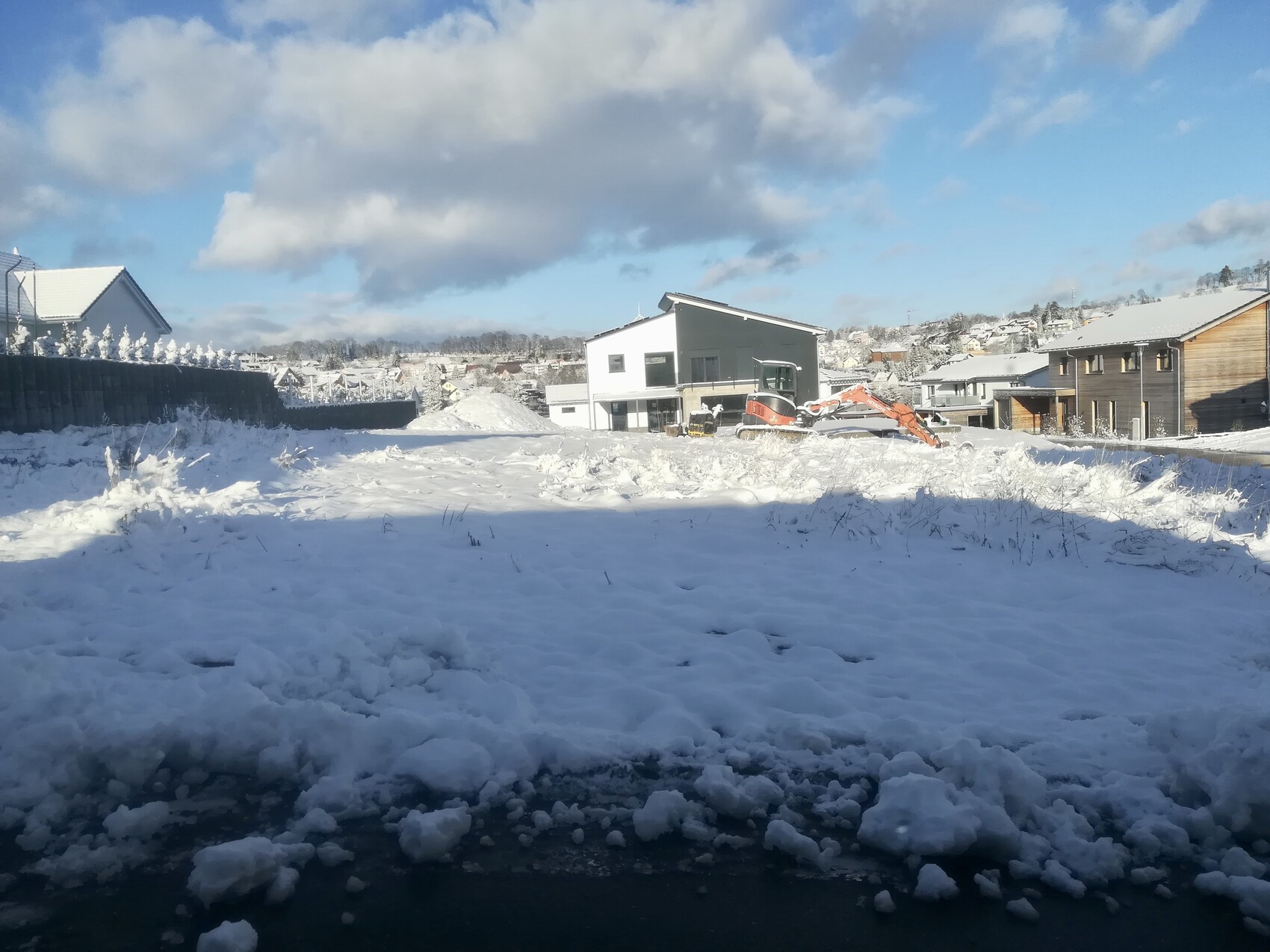 Winterliche Baustelle mit orangefarbenem Bagger vor modernen Häusern in verschneiter Siedlung.