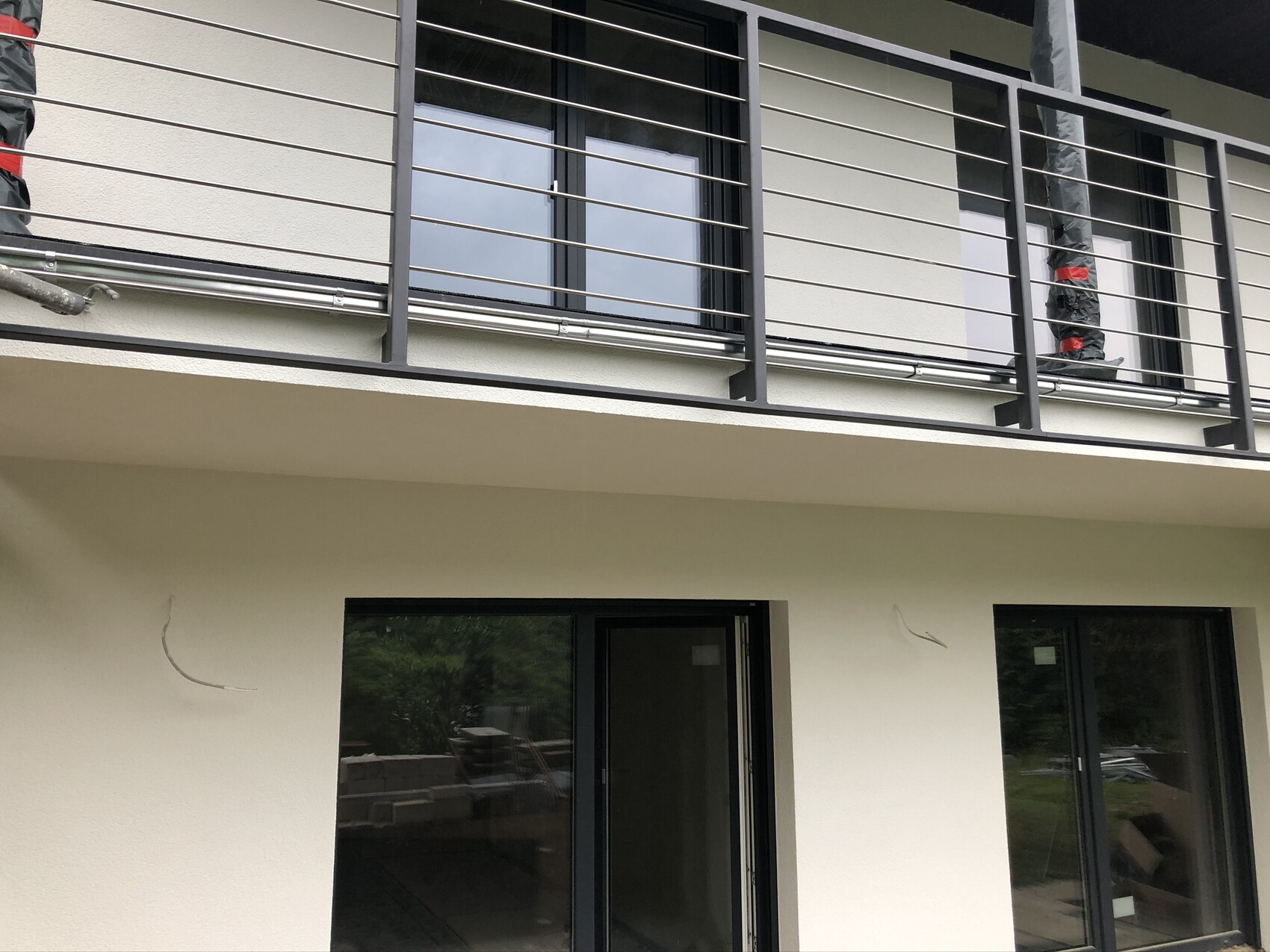 Außenansicht: Balkon mit Metallgeländer und verpacktem Gegenstand; Erdgeschoss mit Glasfenstern.