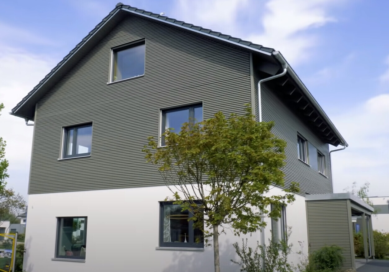 Modernes zweistöckiges Haus mit grauer Fassadenverkleidung, weißem Sockel und Baum im Vorgarten.
