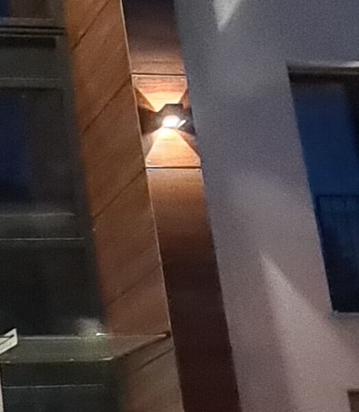 Nächtliche Gebäudeecke mit Wandlampe an brauner Holzverkleidung.