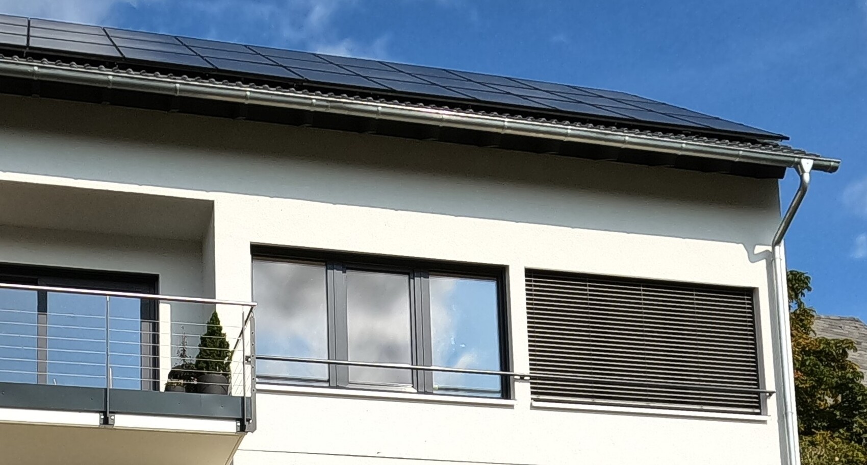 Moderne Hausfassade mit Balkon, großen Fenstern, Rollläden und Solarpanelen auf dem Dach