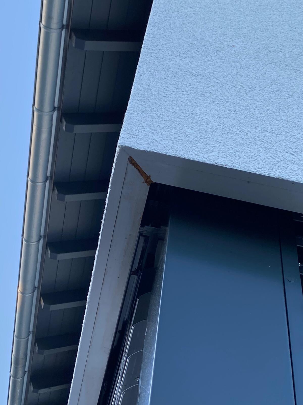 Außenansicht einer Gebäudefassade: blaue Wand, links Abflussrohr, rostige Ecke am Rahmen.
