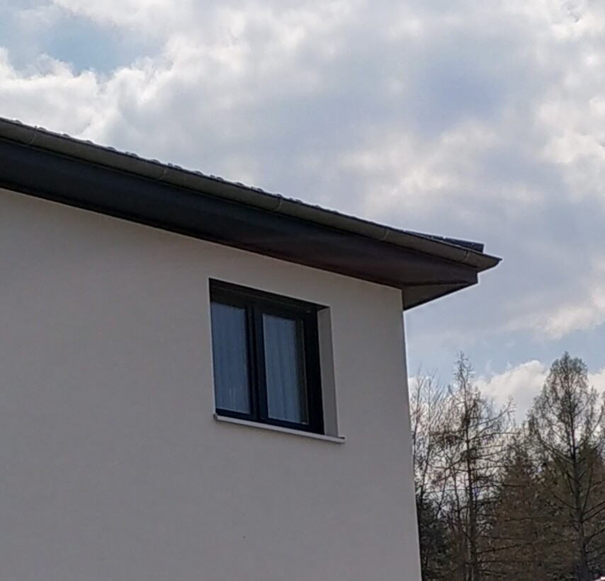 Weiße Hauswand mit dunkel gerahmtem Fenster, schrägem Dach und Bäumen im Hintergrund.