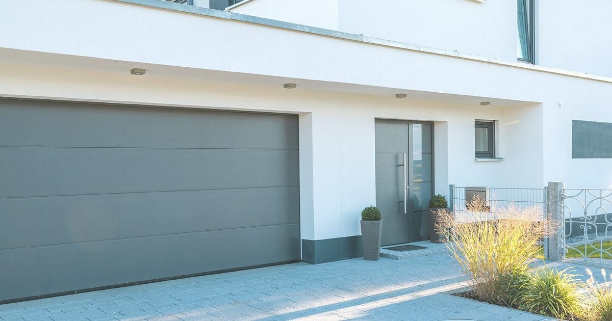 Moderne weiße Hausfassade mit grauer Garage, Eingangstür, Pflanzkübel und Gartenbereich.