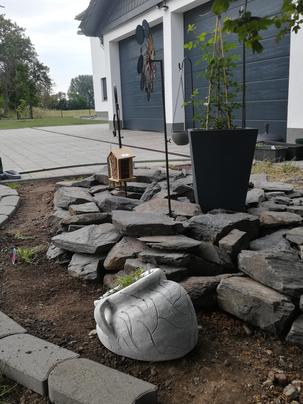 Garten vor modernem Haus mit Steinaufbau, Vogelhaus, Hängekorb und großem Pflanzkübel.