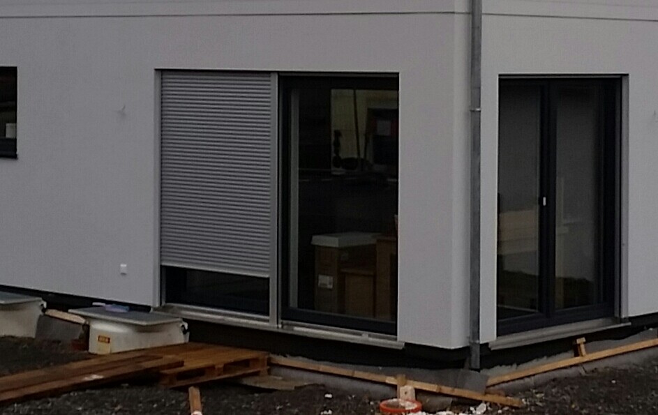 Außenansicht eines modernen Hauses mit Rollladenfenster, Glastür und Bauholzresten.