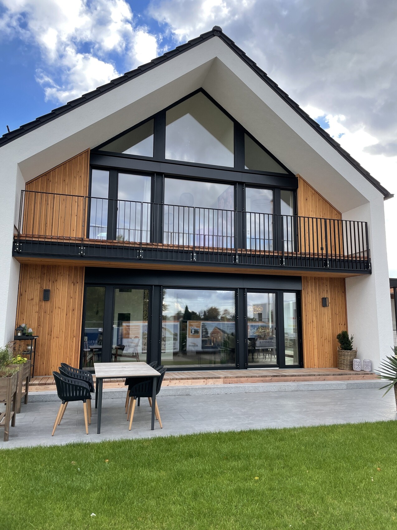 Zweistöckiges Haus mit großer Glasfront, Holzverkleidung, Balkon und Terrasse mit Tisch und Stühlen