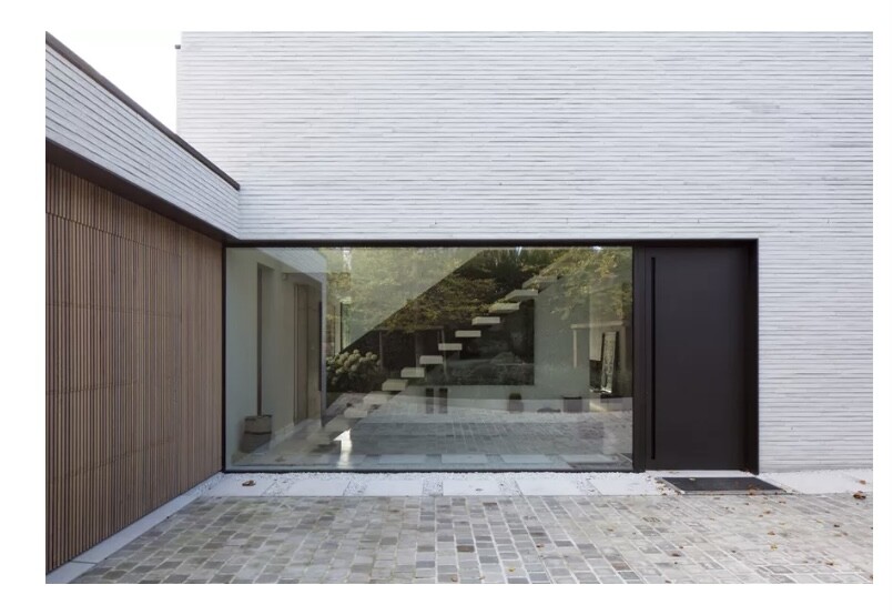 Moderne Hausfassade mit breiter Glasfront, schwebende Treppe innen sichtbar, dunkle Tür rechts.