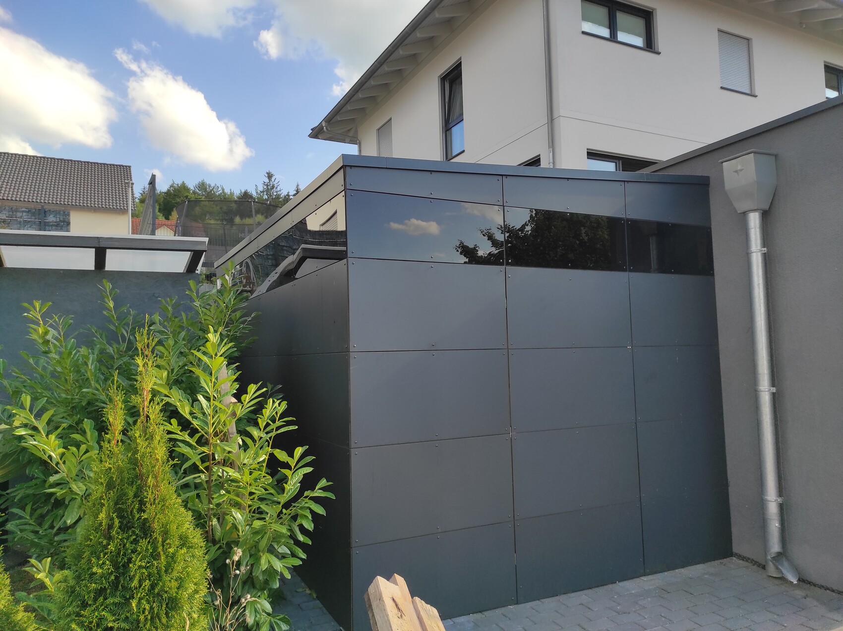 Moderne graue Metallverkleidung eines Anbaus am Wohnhaus, vorn Buschwerk und blauer Himmel.