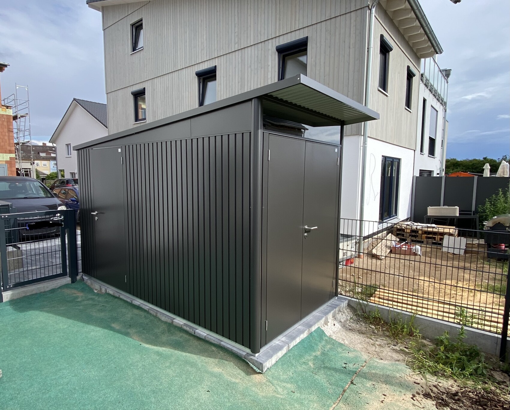 Großer dunkler Metall-Abstellschuppen mit Flachdachüberdachung neben modernem Wohngebäude.