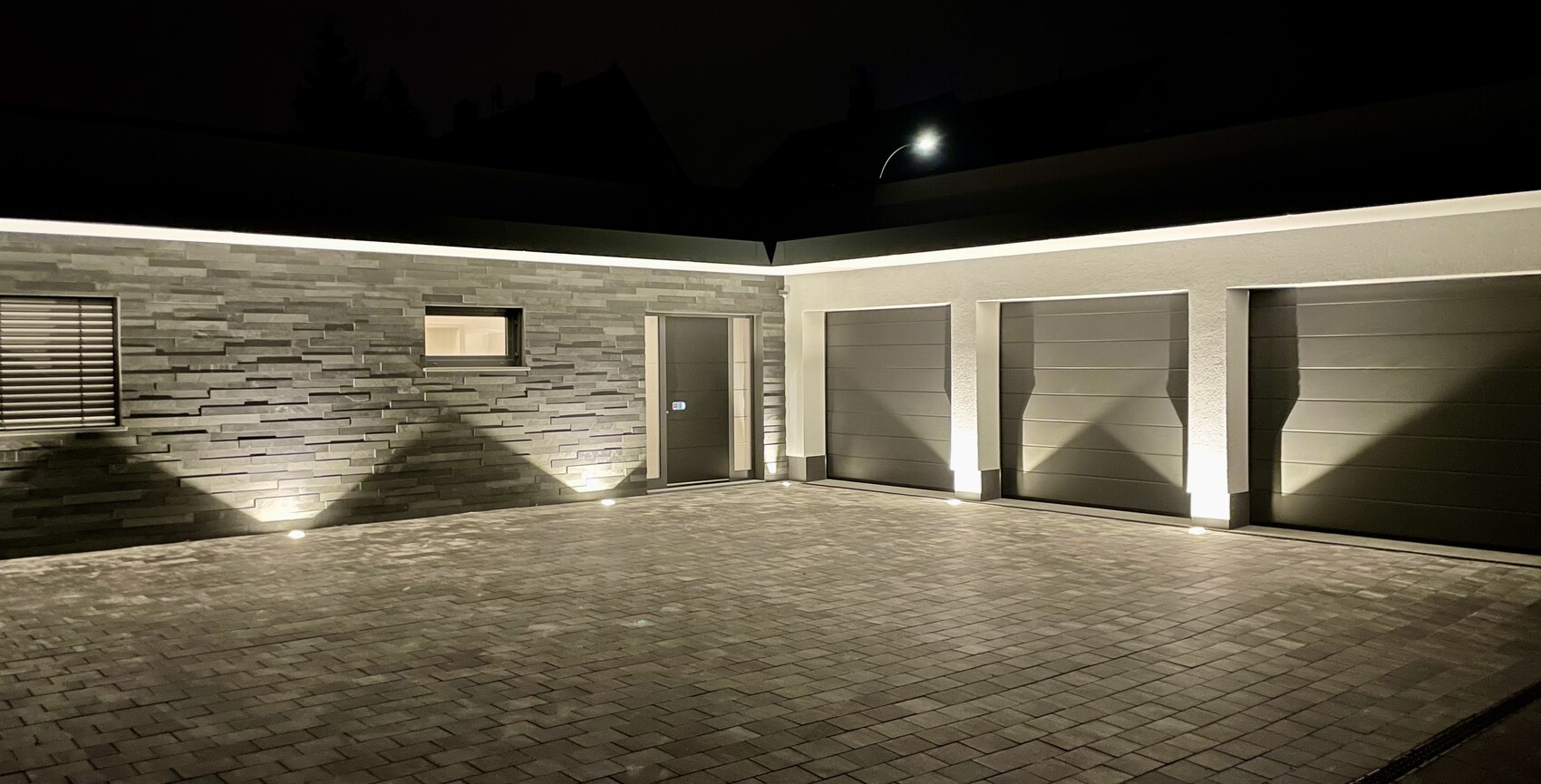Nachtansicht eines modernen Hauses: Steinwand, Glas-Tür, Fenster und drei Garagen mit Beleuchtung.