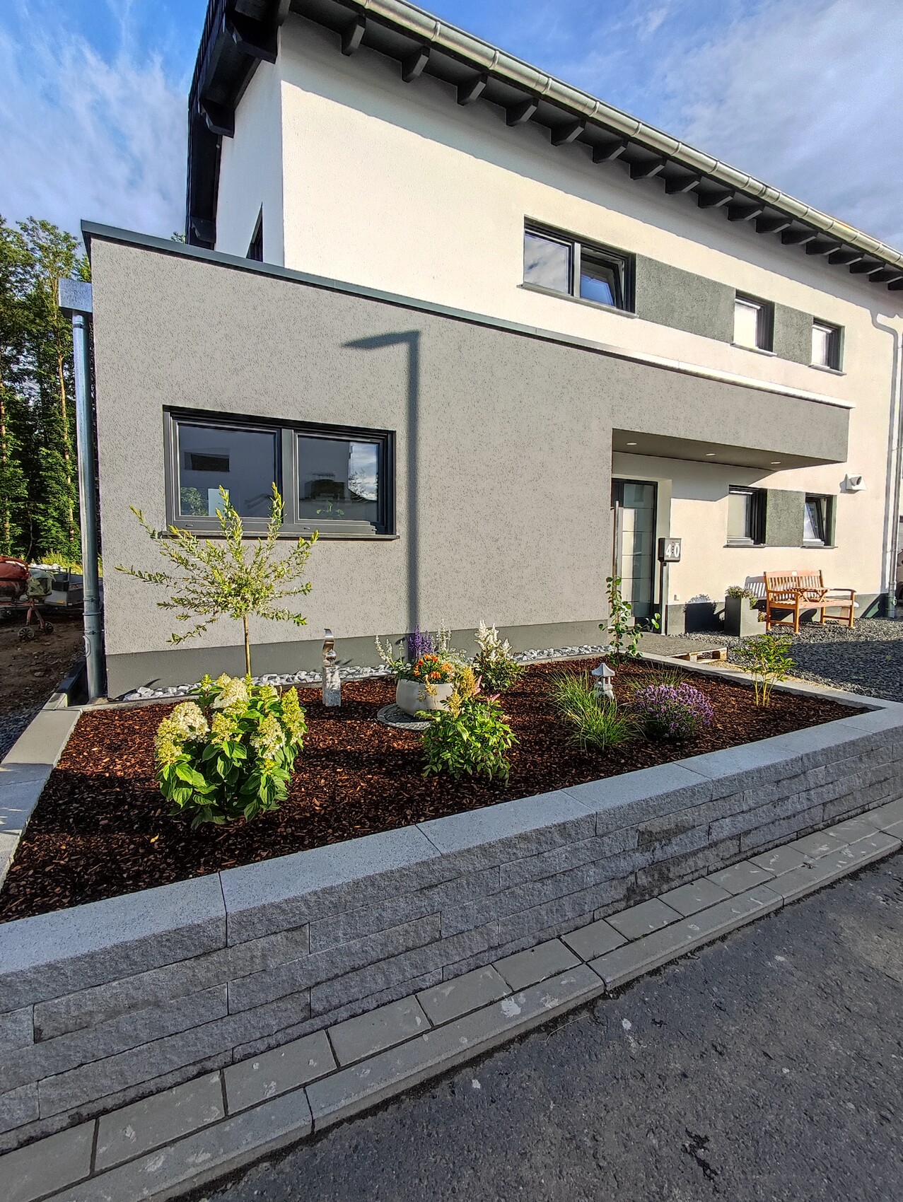 Modernes zweistöckiges Haus mit grauer Fassade; Vorgarten mit Pflanzenbeet und Holzbank.
