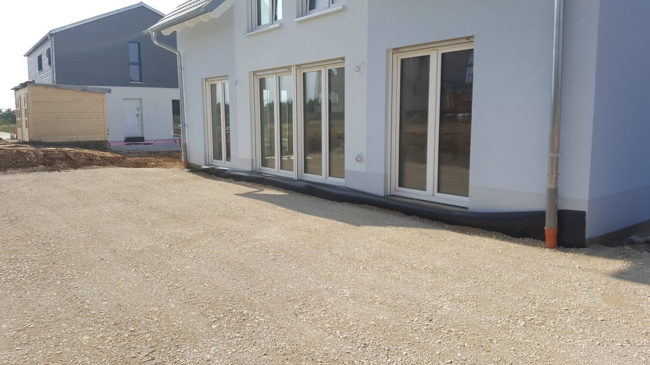 Weiße Hausfront mit großen Glas-Schiebetüren, Kiesboden und Baugebiet im Umfeld