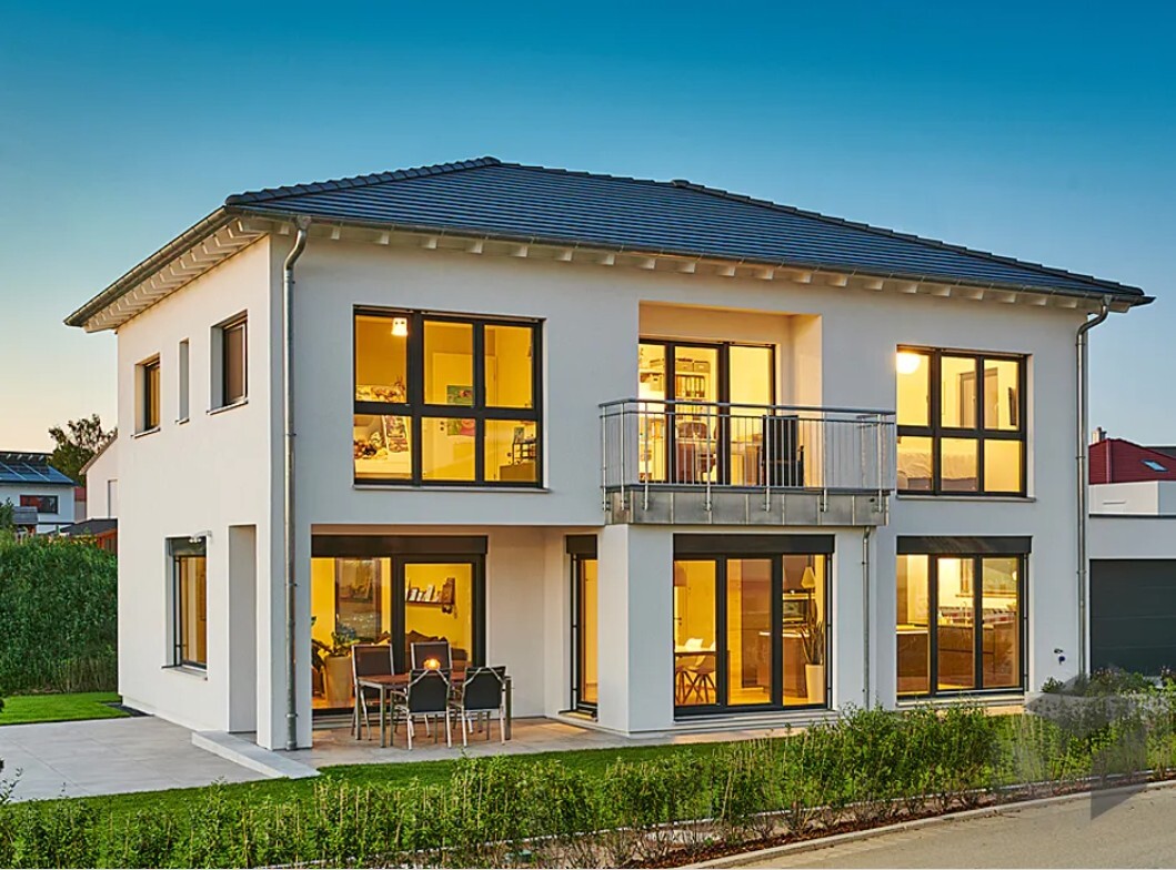 Modern zweistöckiges, helles Haus mit großen Fenstern, Balkon und Terrasse bei Abendlicht.