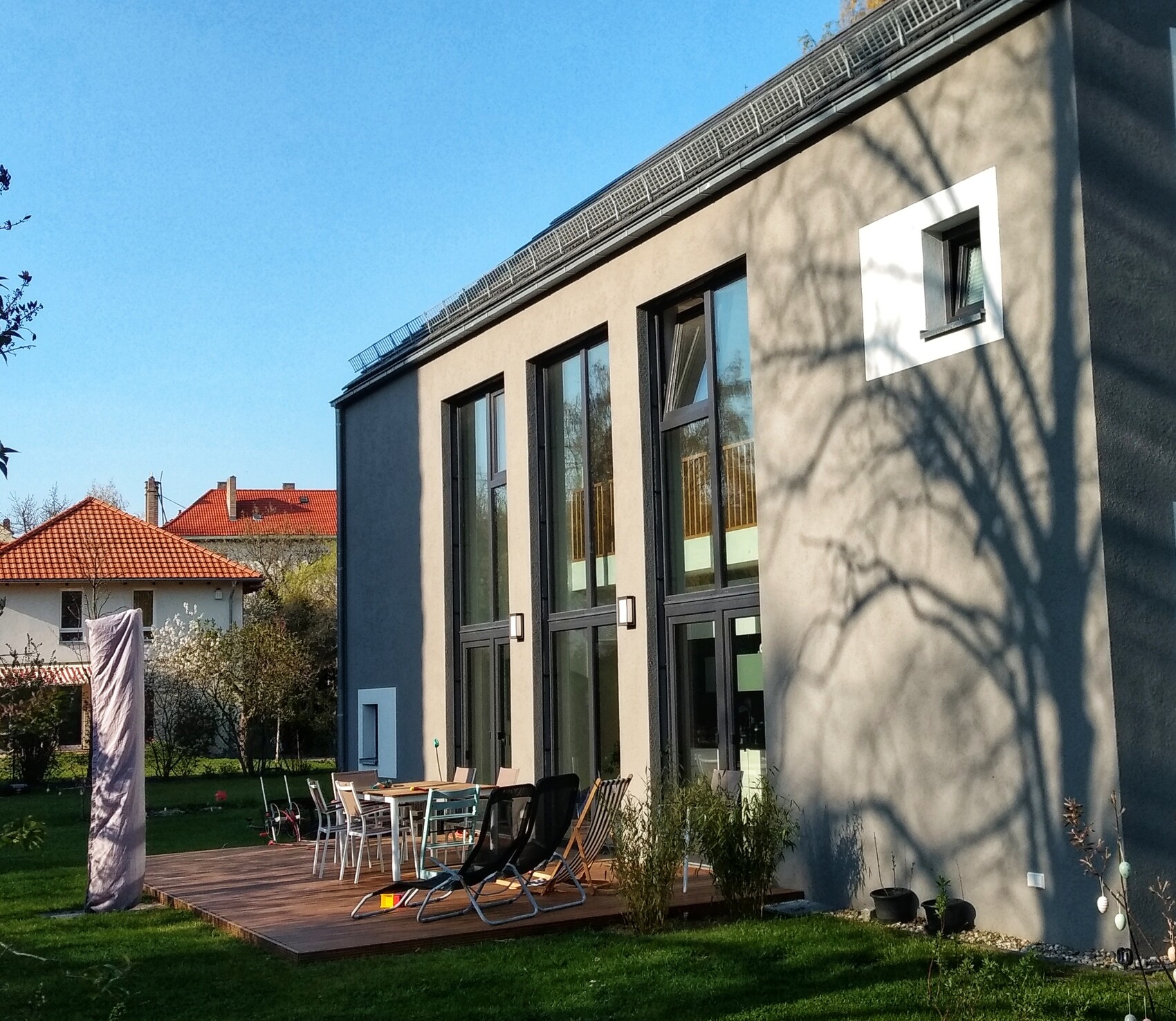 Moderne Gebäudefassade mit großen Fenstern, Holzterrasse, Garten und Baumschatten an der Wand