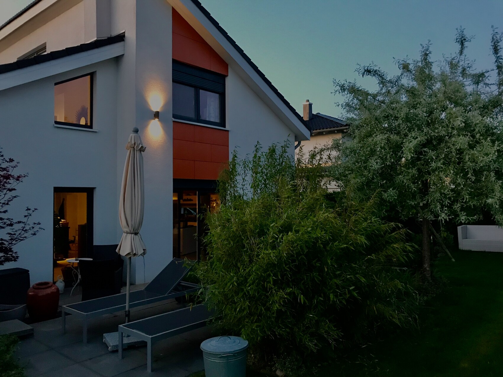 Moderne Hausfassade mit Terrasse, zwei Liegen, geschlossener Sonnenschirm und Garten bei Abendlicht.