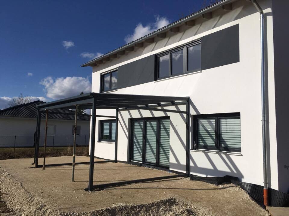 Modernes weißes Haus mit großen Glasfenstern und schwarzer Metall-Pergola im Vorgarten.