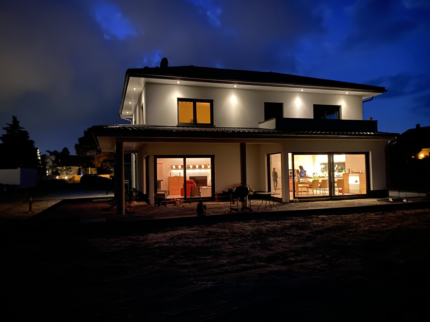 Modernes zweistöckiges Haus bei Nacht, heller Innenraum mit Wohnzimmer und Küche hinter Glasfenstern.