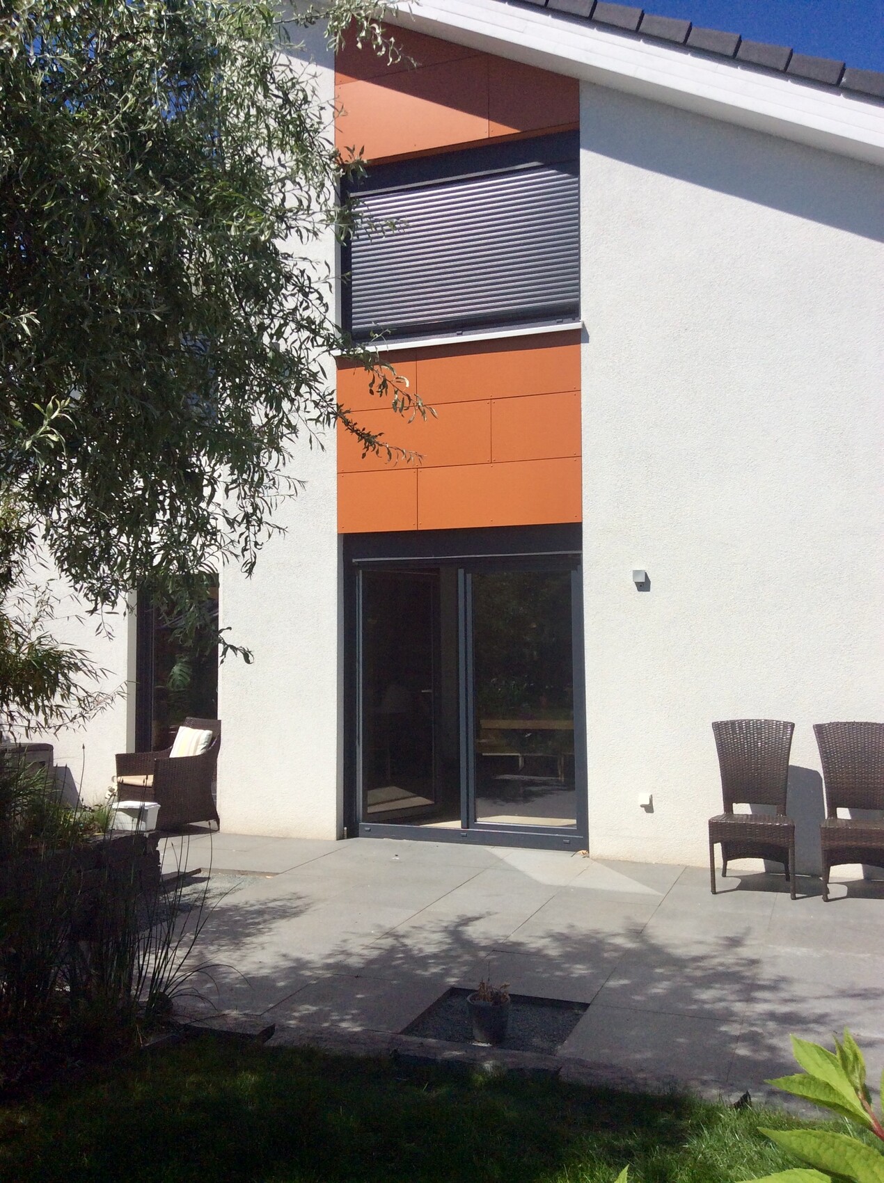 Moderne Hausfassade mit orangefarbenen Paneelen und großen Glas-Schiebetüren Terrasse und Gartenstühle