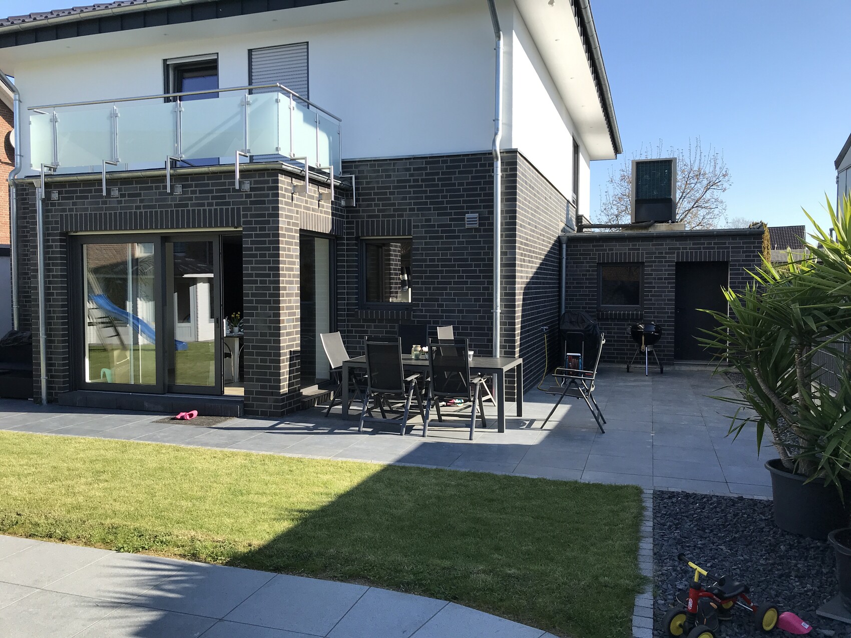Moderne Hausfassade aus dunklem Backstein mit Glasbalkon, Terrasse und Esstischgruppe.