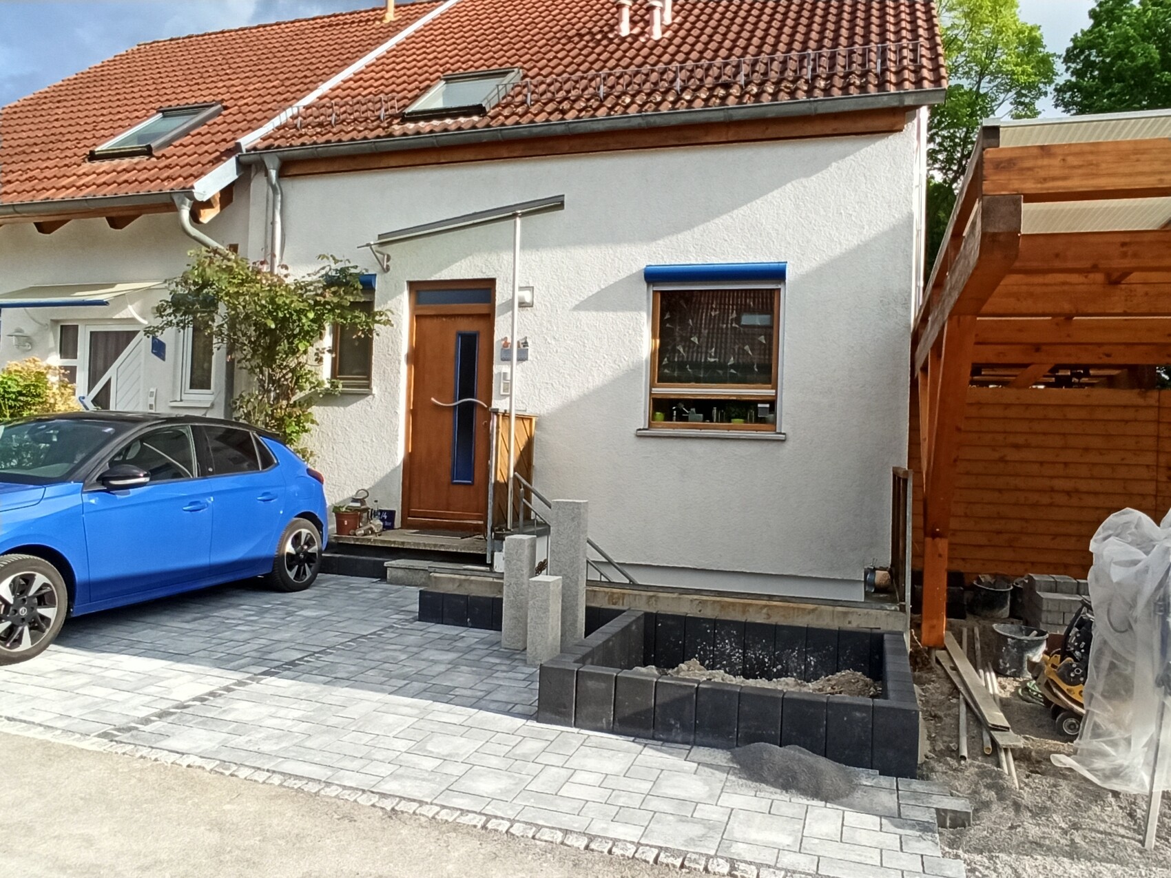Vorderansicht eines Hauses mit rotem Ziegeldach, blauem Auto, Holztür und Baugrube rechts.