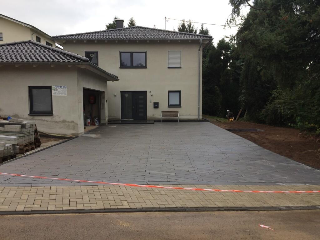 Zweistöckiges Haus mit gepflasterter Einfahrt; links Bauzubehör, rechts Bäume.