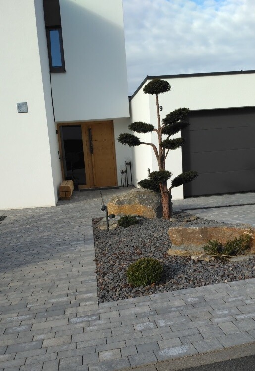 Moderne Hausfront mit Eingangstür, gepflastertem Vorplatz, Garage und Garten.
