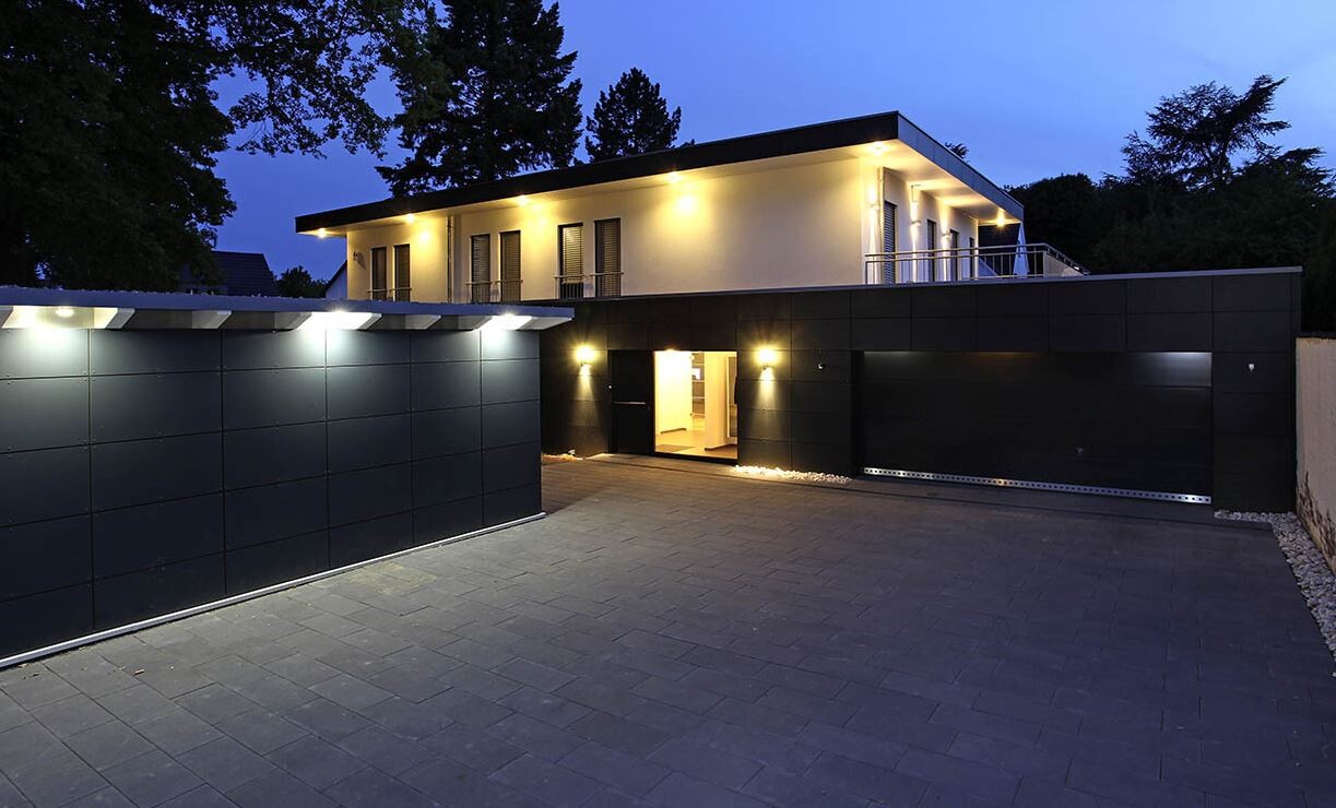 Moderne Villa mit schwarzer Fassade, beleuchtetem Eingang und großer Garage bei Nacht.