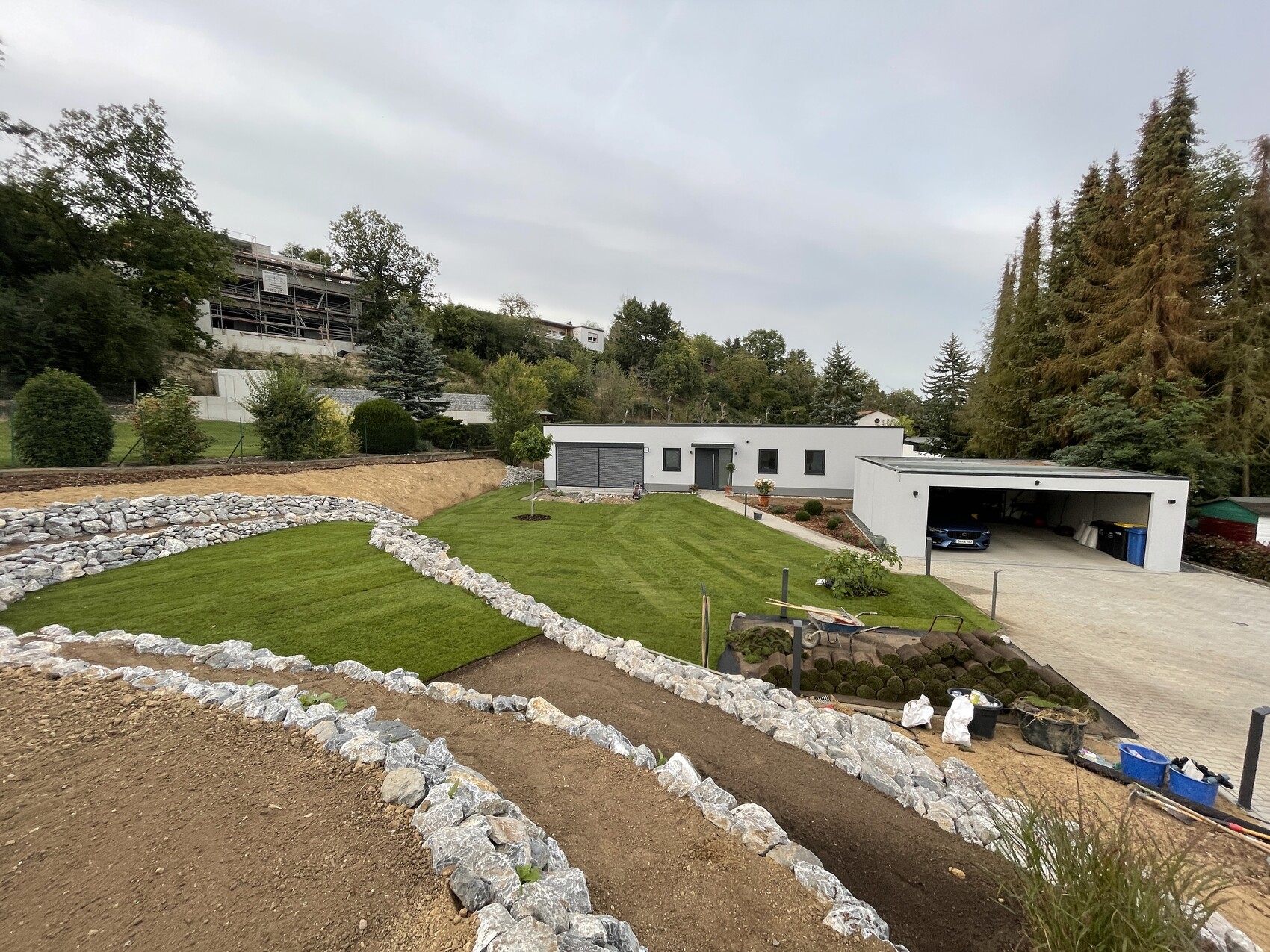 Moderne Hausanlage mit Garage, grünem Rasen und steinernen Terrassen; Bauarbeiten dahinter.
