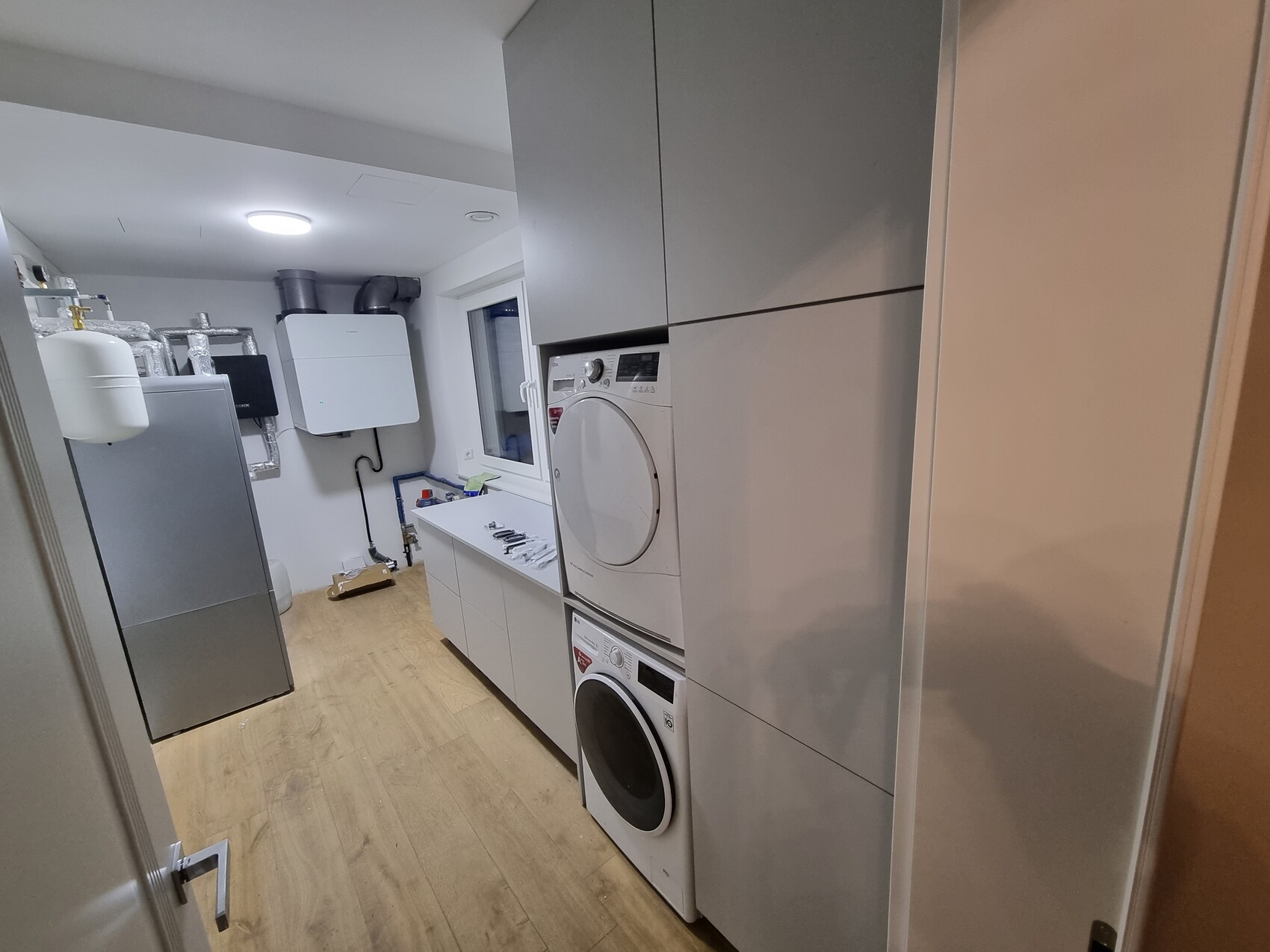 Waschraum mit Waschmaschine unten, Trockner oben, grauen Schränken und Fenster.