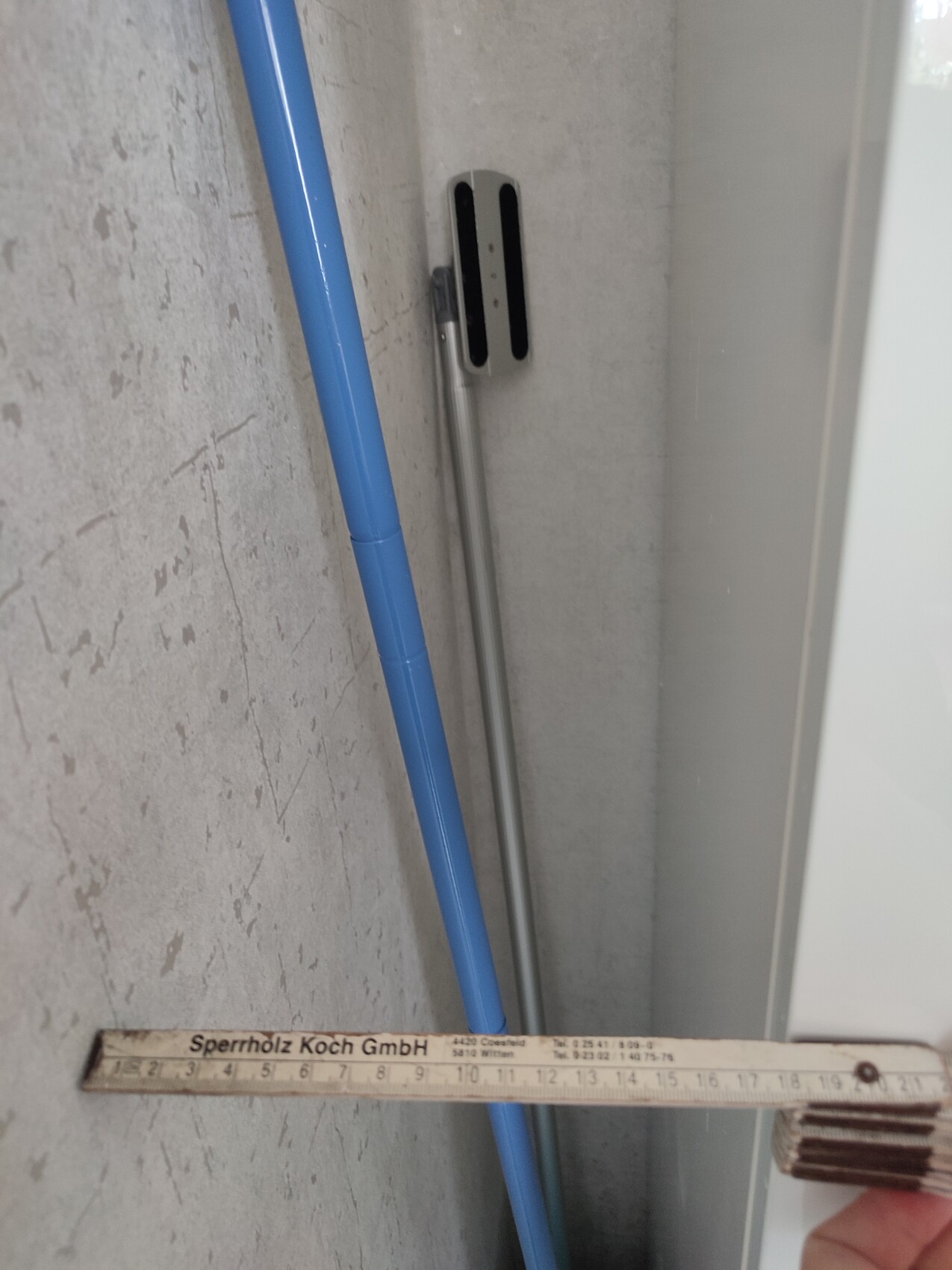 Blaue Reinigungsstange lehnt an grauer Wand; im Vordergrund liegt ein Holz-Zollstock.