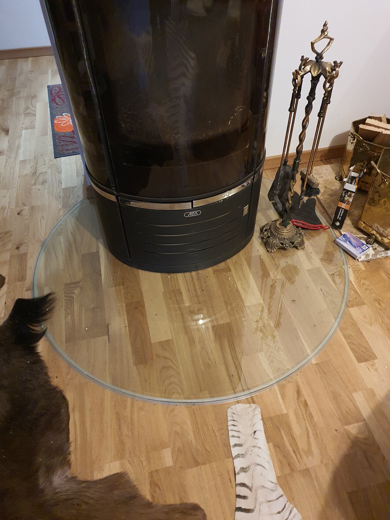 Runder Glasring um schwarzen Kaminofen; Werkzeug und Holz daneben.