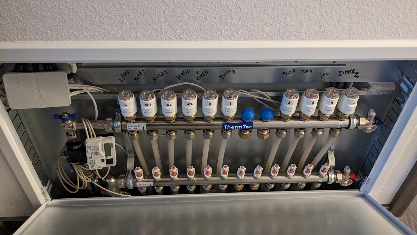 Heizungs-Verteiler mit vielen Thermostatventilen, Rohren und Kennzeichnungen im Gehäuse.