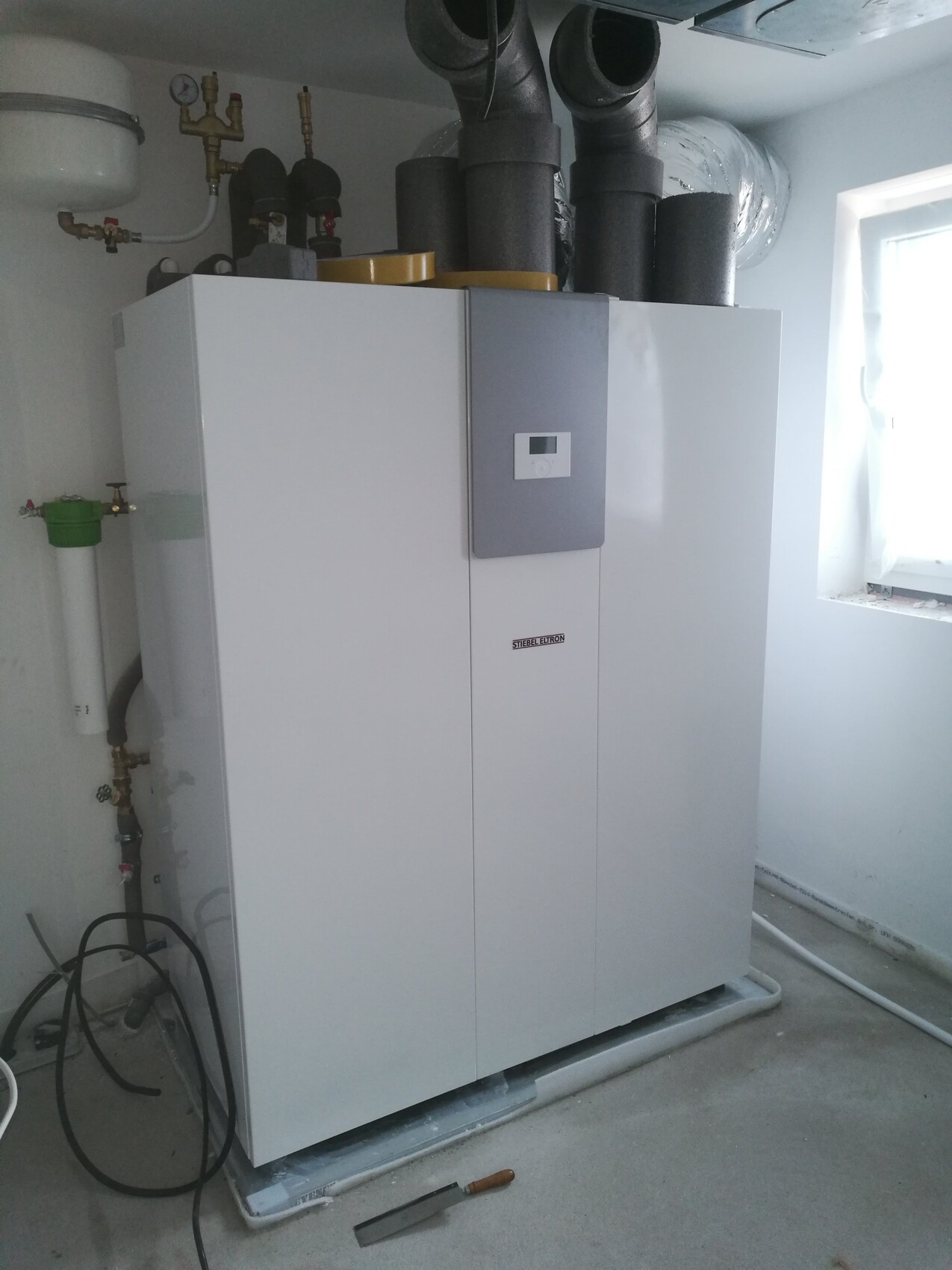 Weiße Heizungsanlage/Boiler im Keller mit Display, Rohren und Dämmung am oberen Bereich.