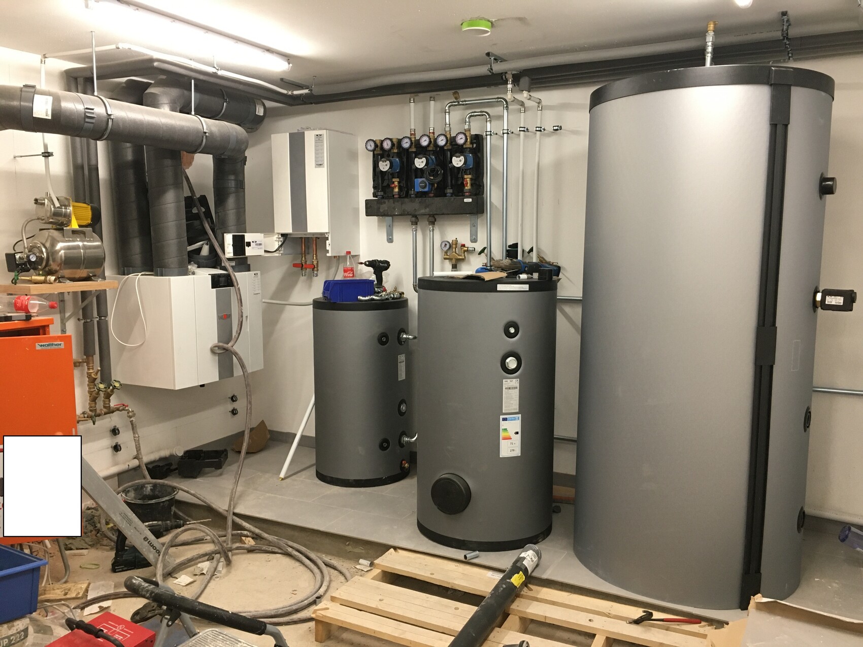 Großer Heizungsraum mit mehreren grauen Speichertanks, Rohren, Messgeräten und Pumpen.