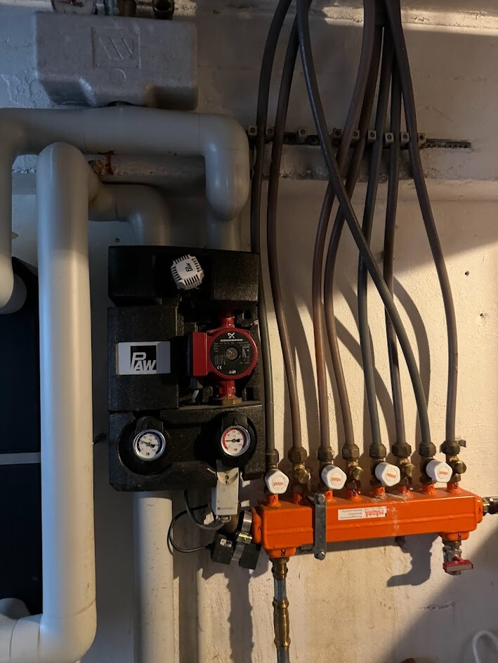 Heizungsstation mit Pumpe, Druckanzeigen und orangefarbenem Verteiler mit Ventilen.