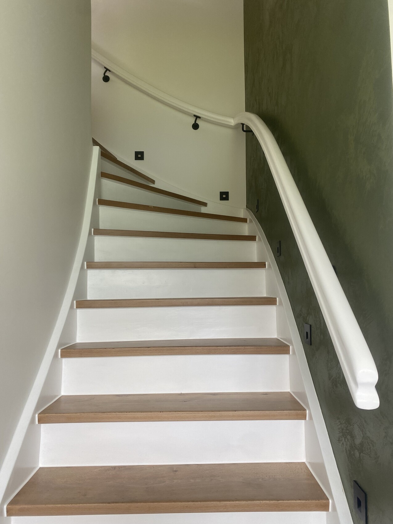 Weiße Treppe mit Holzstufen, rechts gebogener Handlauf neben grüner Wand.