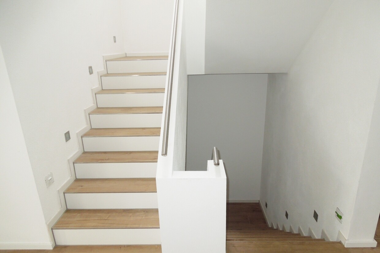 Weiße Treppe mit Holzstufen und Metallhandlauf in hellem, modernem Treppenhaus.