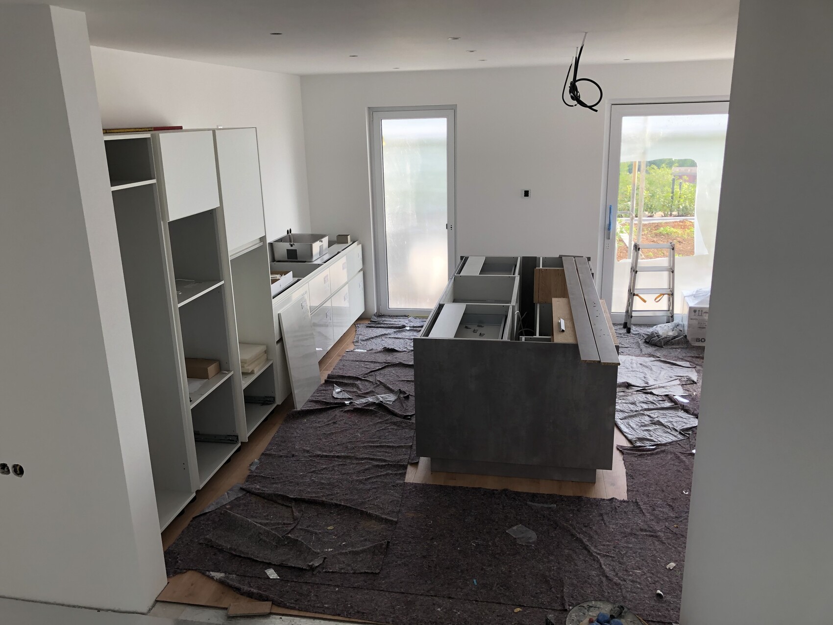 Küche im Bau: Insel in Mitte, weiße Oberschränke, Bodenschutzdecke, Leiter im Raum.