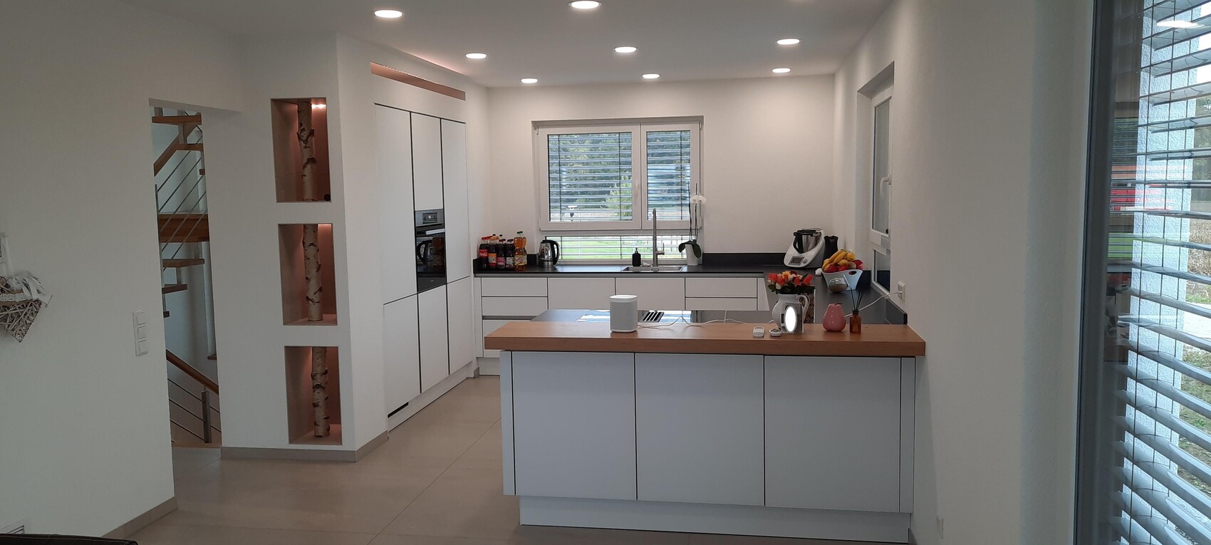 Moderne weiße Küche mit Insel aus Holz, integrierten Geräten, Obstkorb und Fenster mit Jalousien.