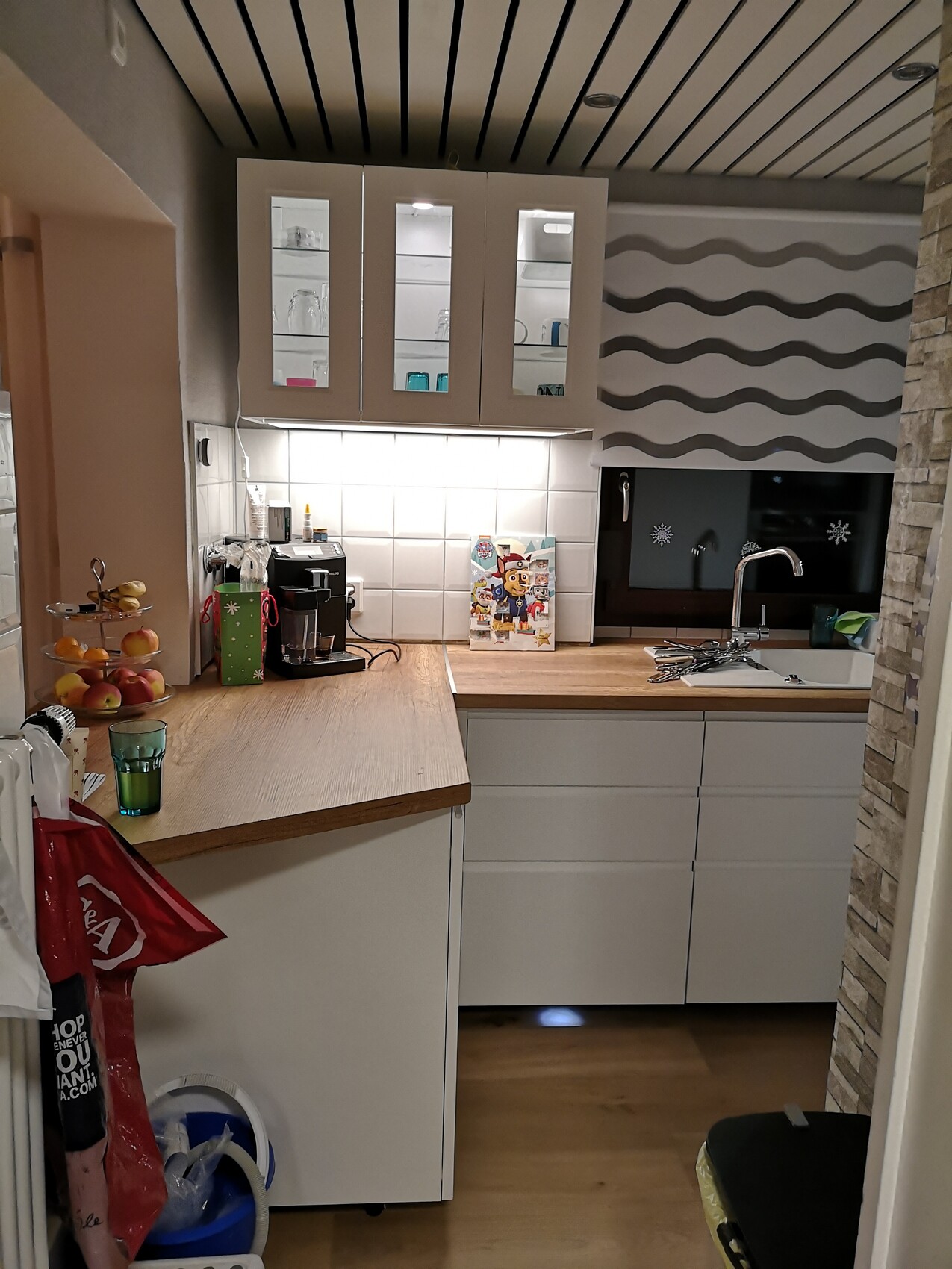 Küche mit weißen Schränken, Gläser in Vitrinen, Kaffeemaschine, Obstkorb, Spüle, Wellenwand.