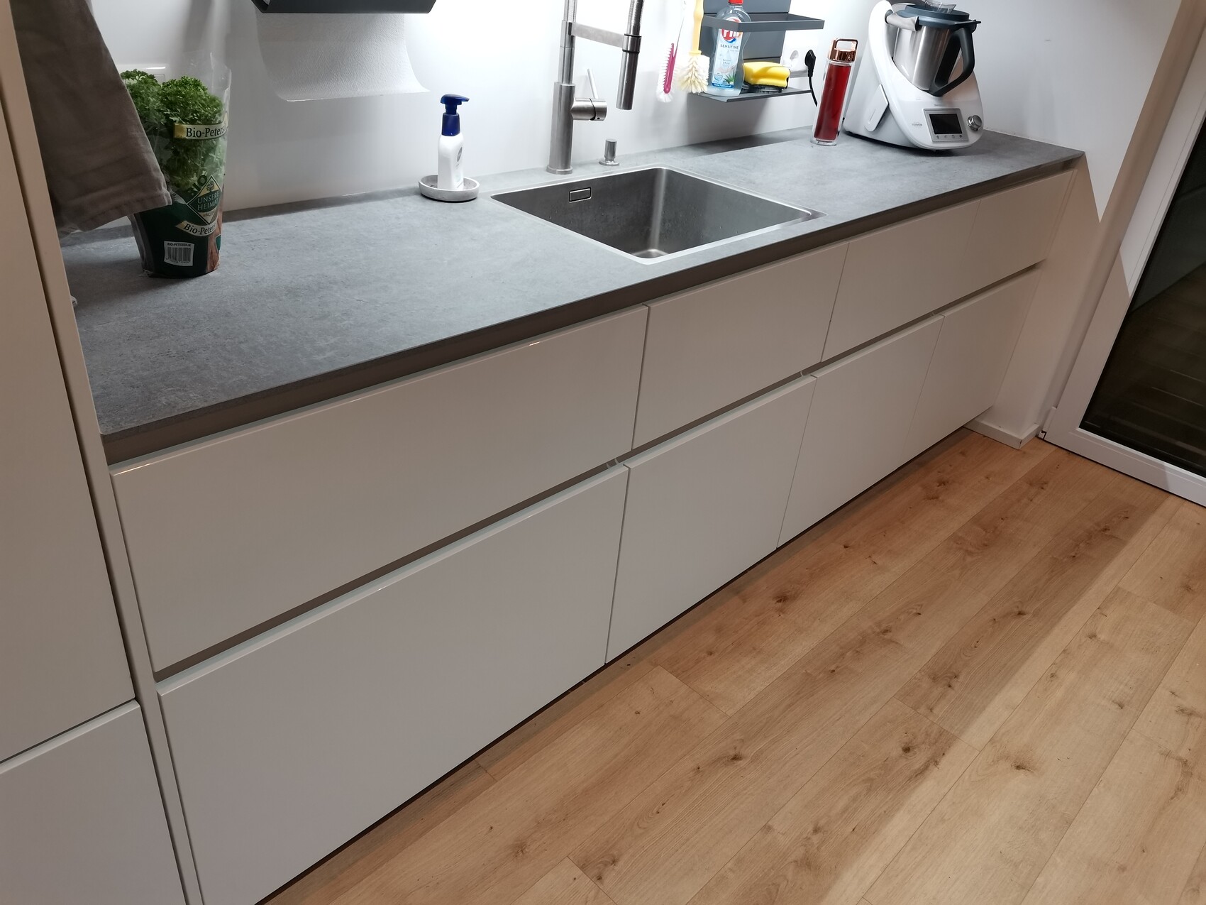 Moderne Küchenzeile mit grauer Arbeitsplatte, Spüle, Wasserhahn, Küchenmaschine und Holzfußboden.