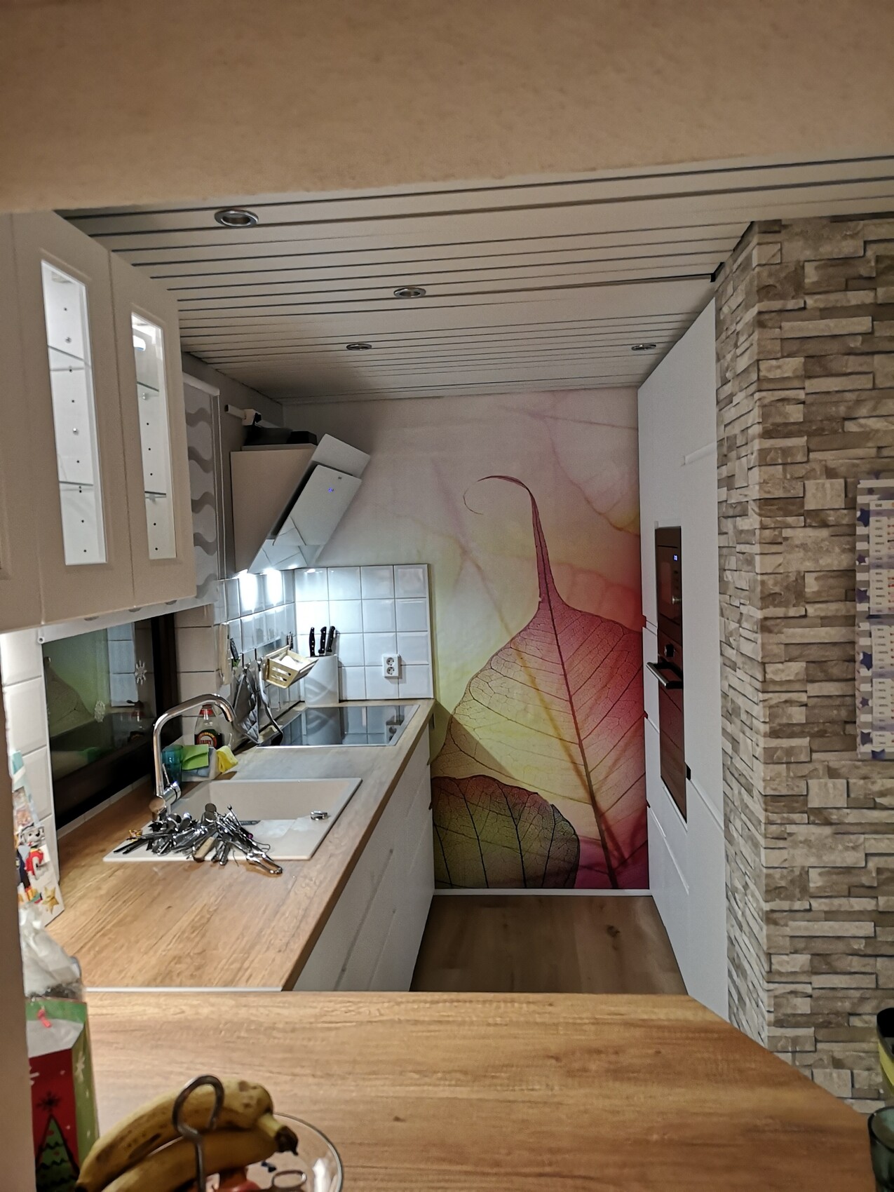 Küche mit Holzarbeitsplatte, weißen Schränken, Spüle und farbigem Blatt-Wandbild.