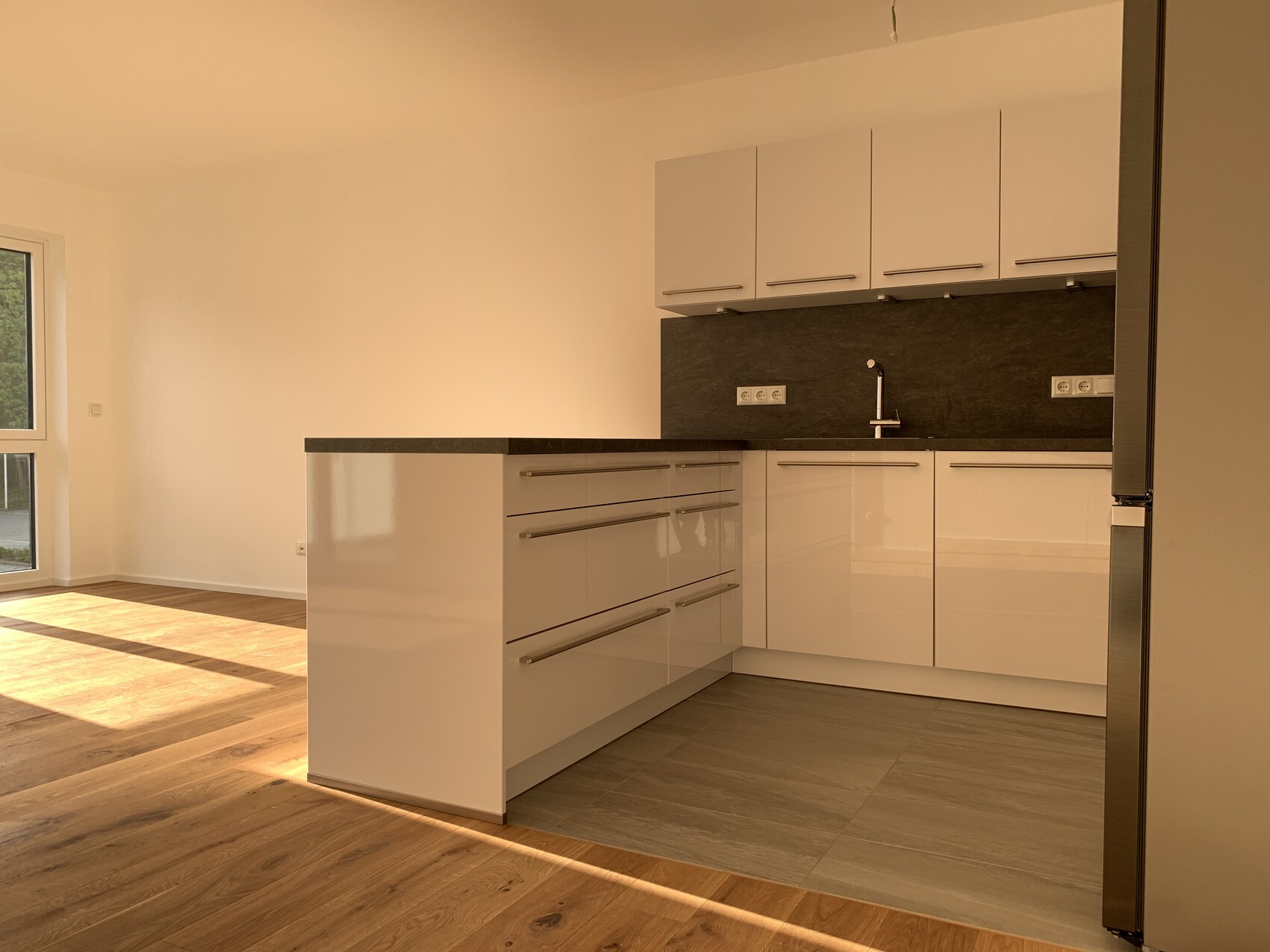 Moderne weiße Küche mit dunkler Steinplatte, Spüle und Fenster, sonnendurchflutet.