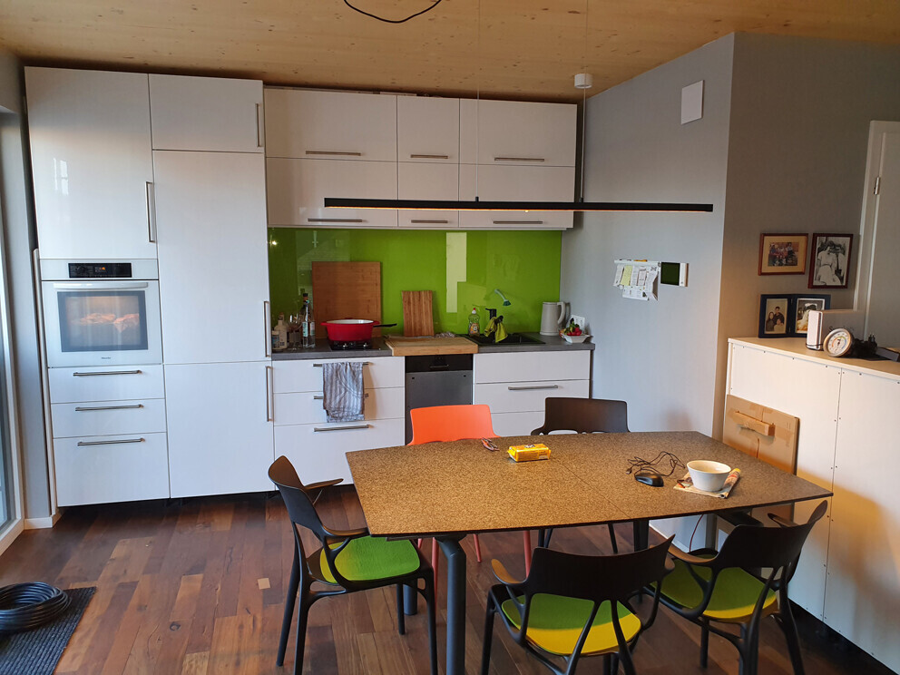 Moderne Küche mit weißen Fronten, grüner Fliesenrückwand, Esstisch und bunten Stühlen.