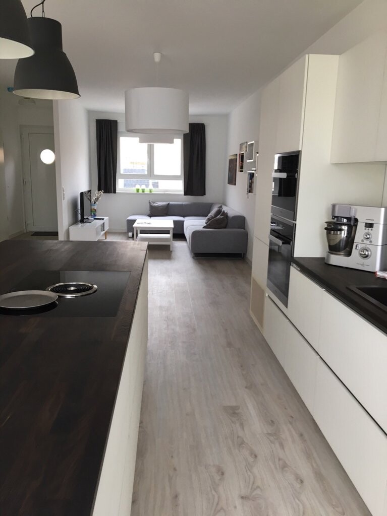 Offene Küche mit dunkler Insel-Arbeitsplatte, weißen Schränken; Wohnzimmer mit grauem Sofa.