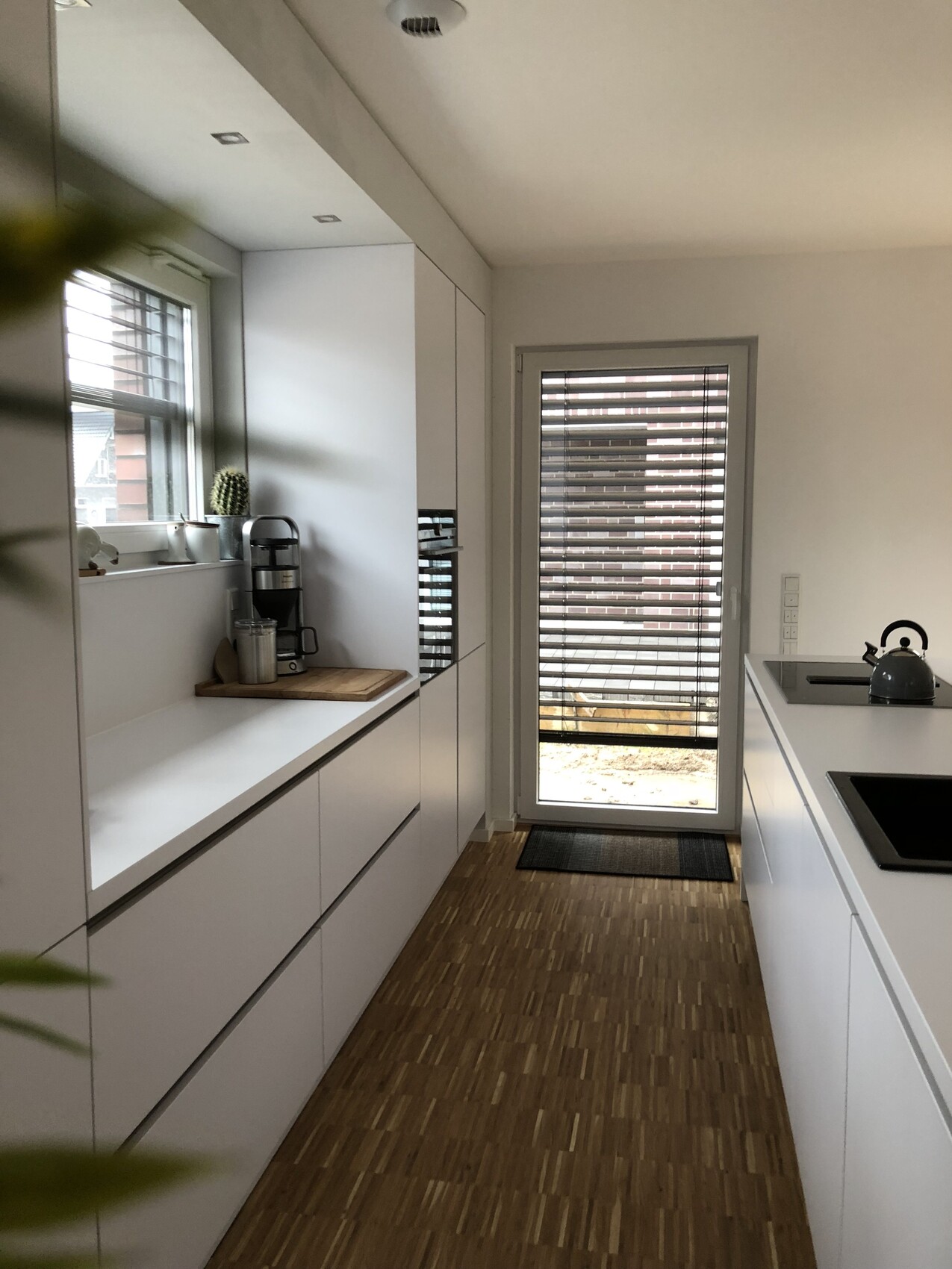 Moderne weiße Küche mit langen Schränken, Holzboden, Kaffeemaschine, Schneidbrett, Tür mit Jalousien