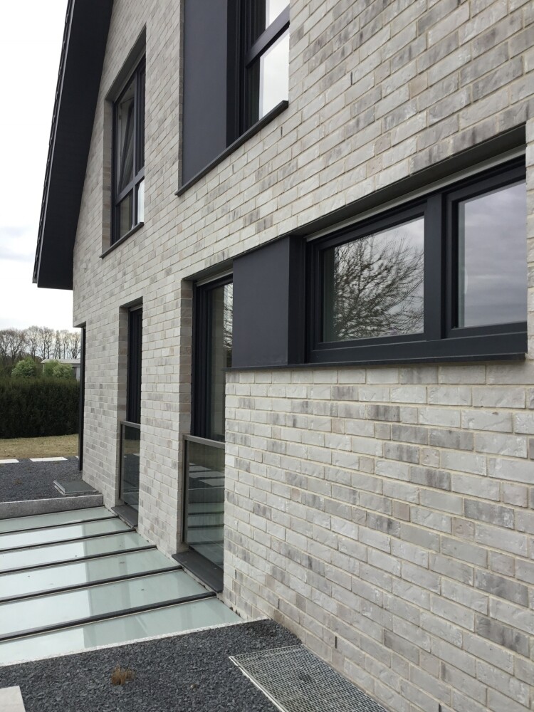 Moderne zweigeschossige Hausfassade aus hellgrauen Ziegeln mit schwarzen Fensterrahmen.